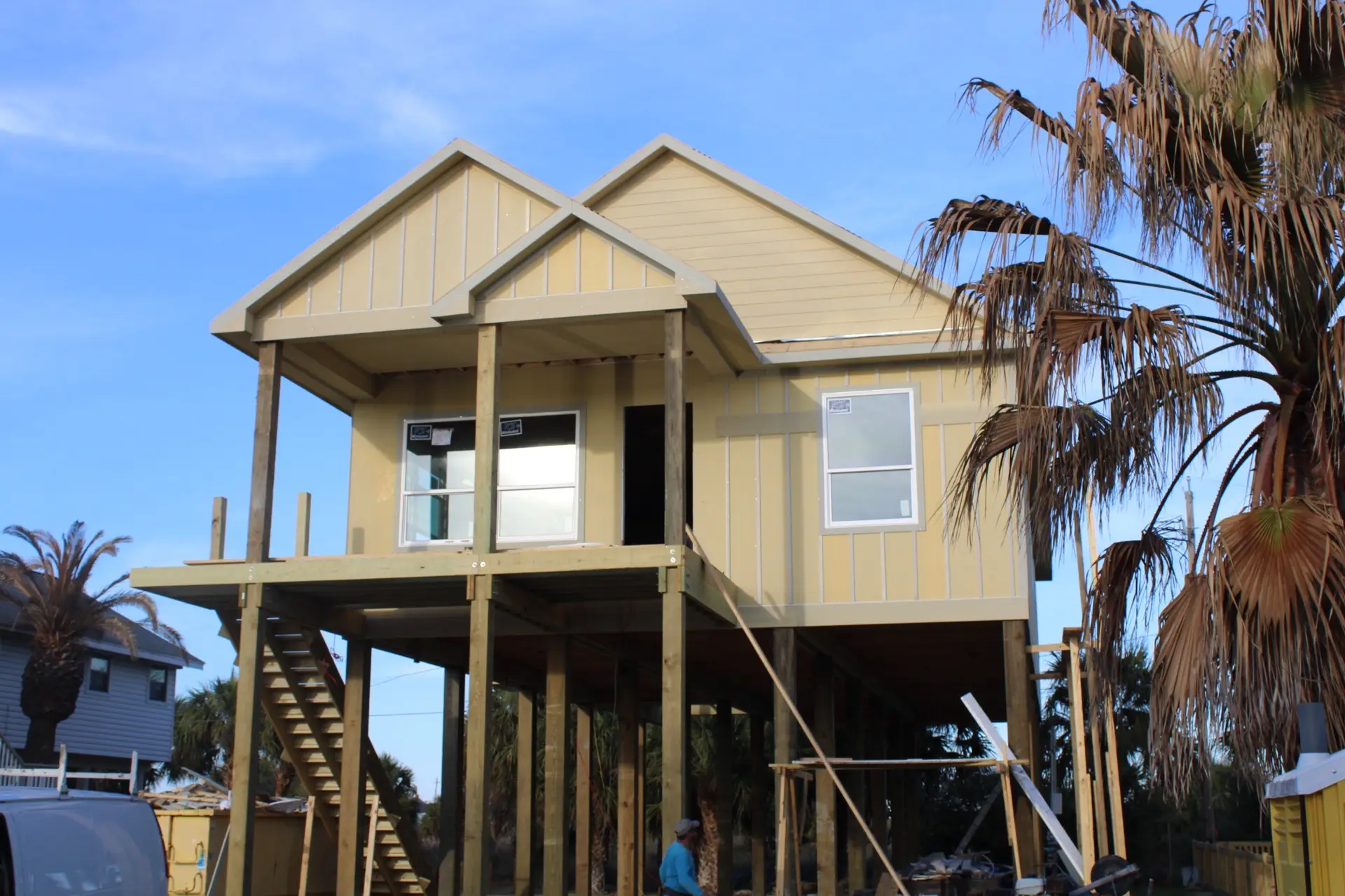 Front exterior of 27 Laguna on Tiki Island featuring warm tan and yellow board-and-batten siding raised on stilts with palm tree landscaping and classic Gulf Coast charm