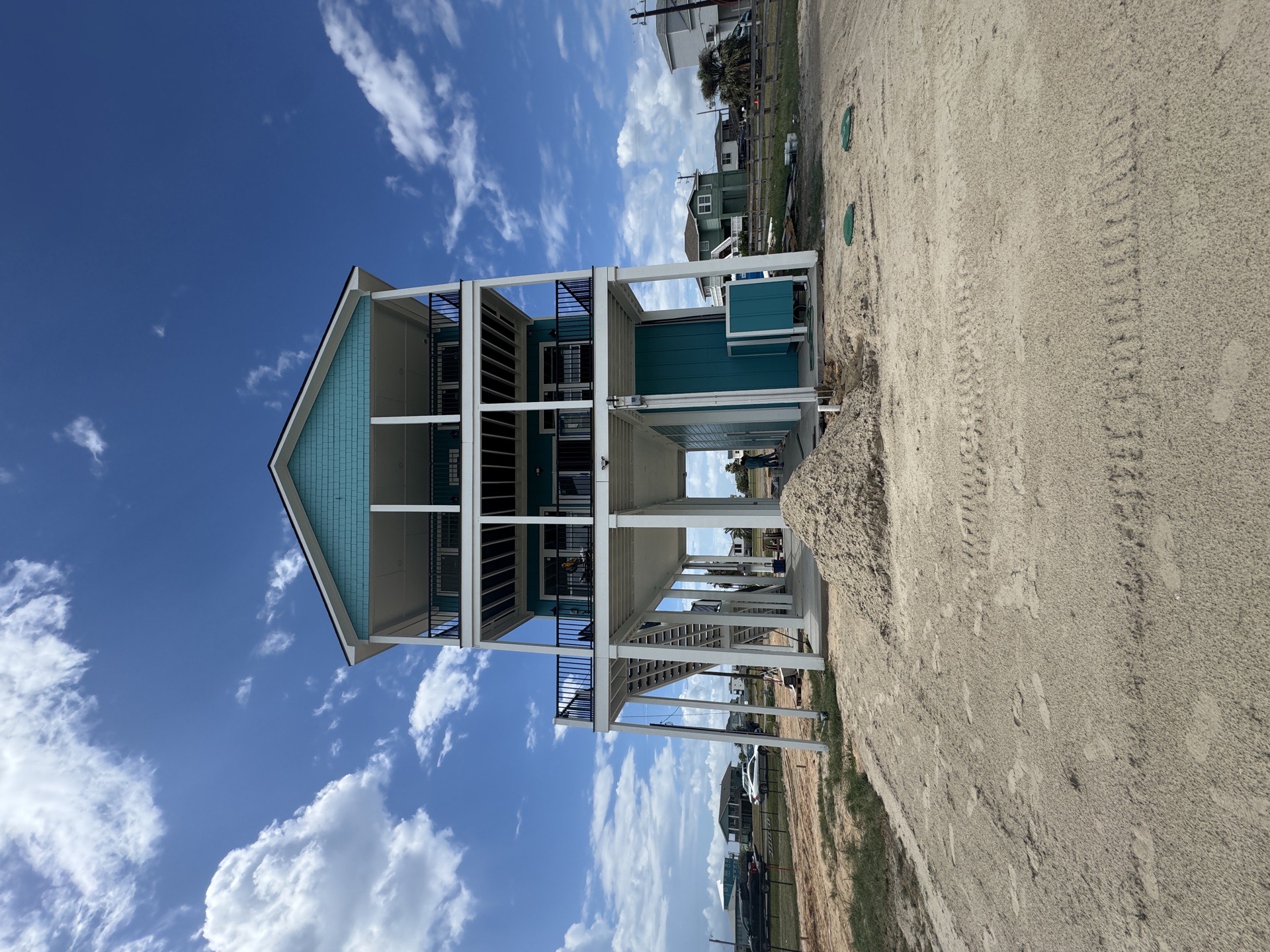 Front exterior of 2nd Street custom home in Galveston featuring striking teal and turquoise board-and-batten siding with white trim, two levels of covered porches with white railings, on a sandy lot near the beach