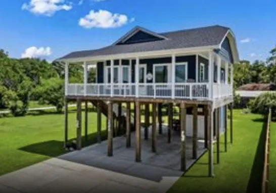 Exterior of the 5th St Seabrook cottage elevated on pilings, with navy blue roof, white railing, and a shaded ground level below