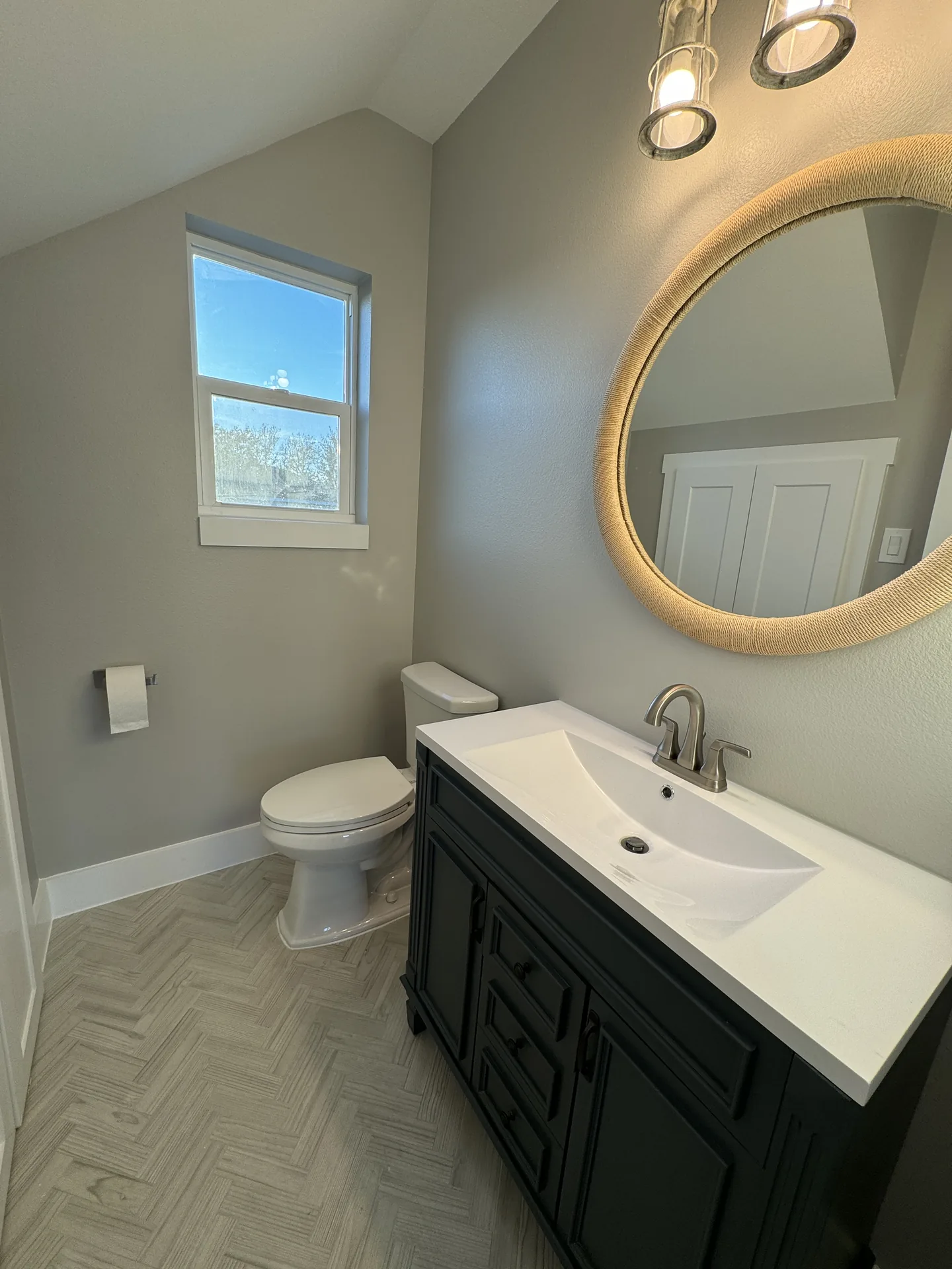 Powder bath at Galleon with a round gold-framed mirror, dark vanity, and herringbone tile flooring