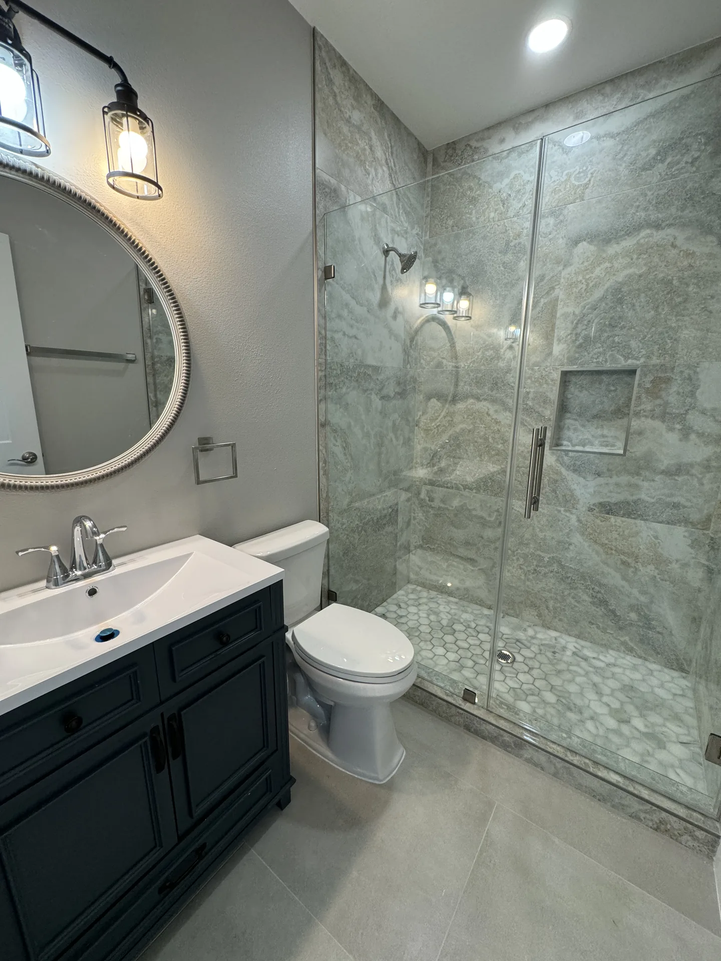 Guest bath at Galleon with a round mirror, navy vanity, and marble-look shower behind glass