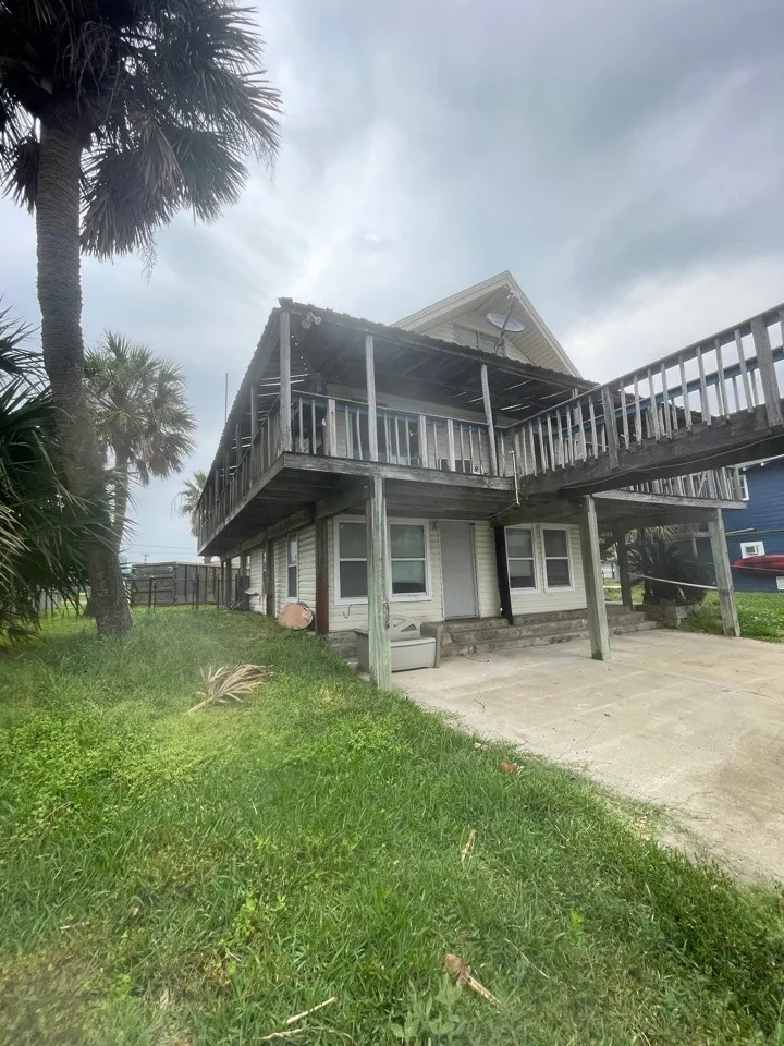 Before: original exterior of the Jamaica Beach A-frame with weathered siding, stacked decks, and overgrown yard