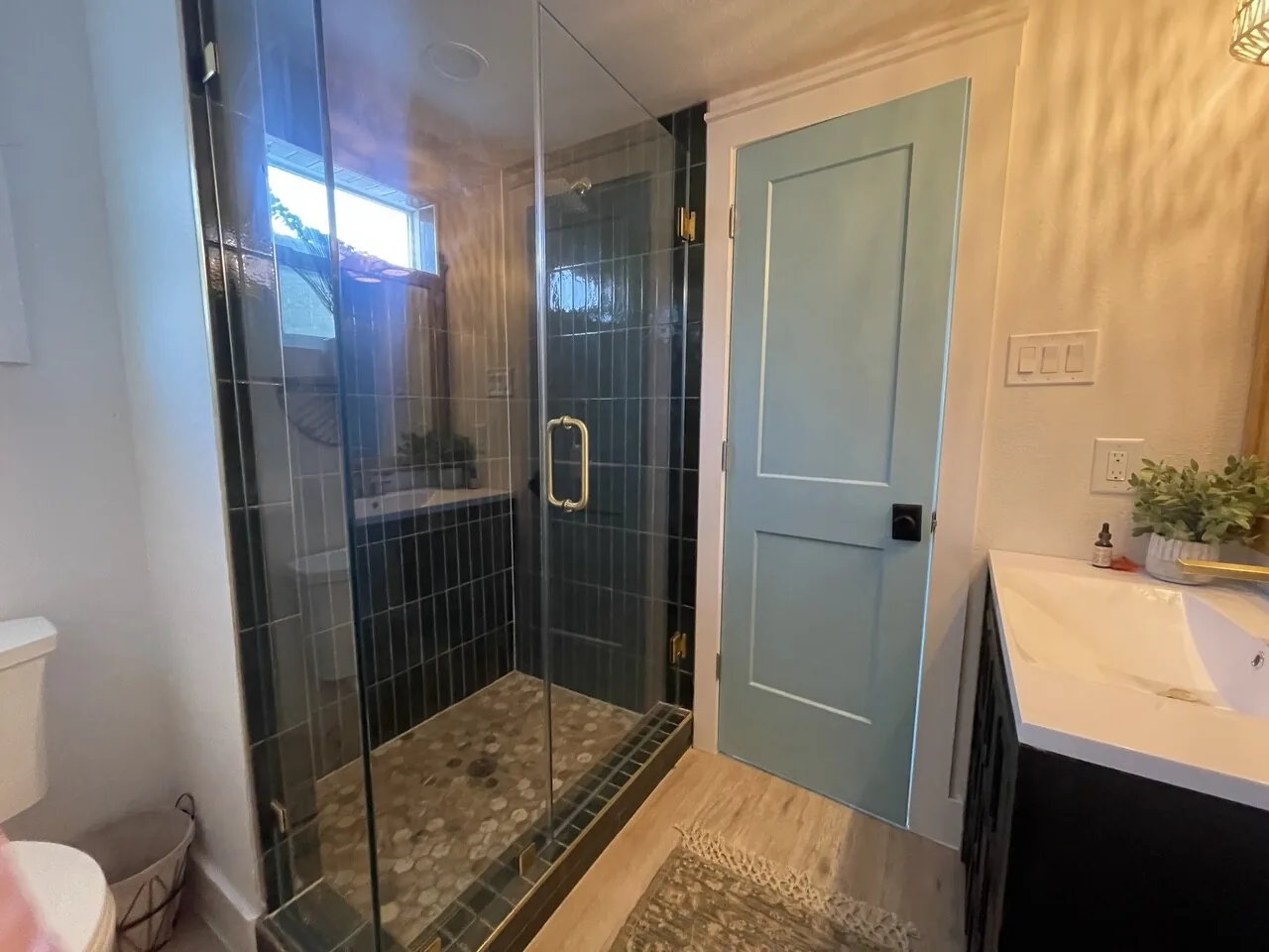 After: renovated walk-in shower with frameless glass, wood-look tile, and a teal painted door